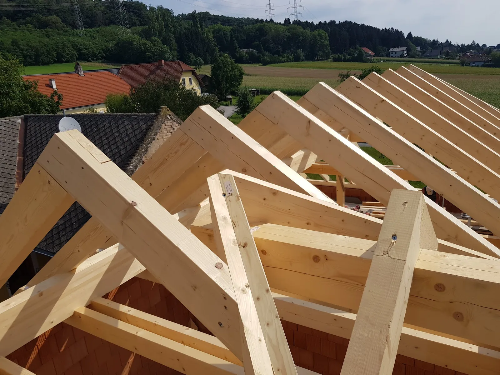 Blick von oben auf einen fertig aufgestellten Dachstuhl über einem Ziegelrohbau, dahinter Ortsrand und Felder